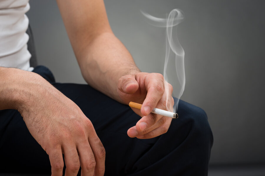 Nahaufnahme einer sitzenden Person mit den Händen auf den eigenen Knien, die eine brennende Zigarette zwischen den Fingern hält, mit sichtbar aufsteigendem Rauch. 