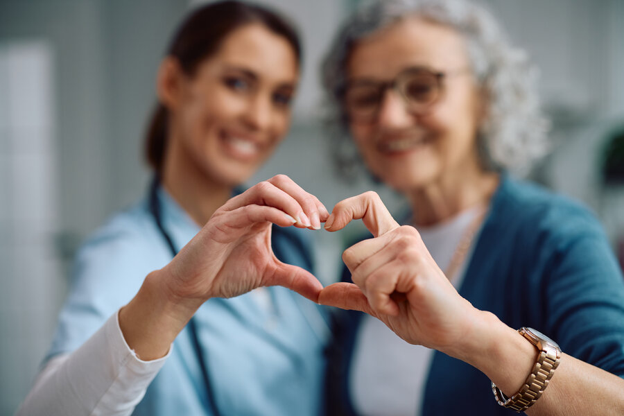 A young doctor in blue scrubs and an elderly patient with grey hair and glasses form a heart shape together with their hands in the foreground, both smiling softly out of focus in the background.