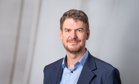 A middle-aged man in a light blue shirt and a navy blazer against a grey background.