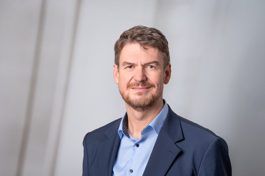 Ein mittelalter Mann in hellblauem Hemd und dunkelblauem Sacko vor grauem Hintergrund.