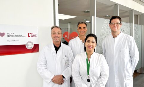 Gruppenfoto des TRIC-I-HF-Studienteams vor einem DZHK-Schild (Deutsches Zentrum für Herz-Kreislauf-Forschung), Standort München, in weißen Kitteln: drei Männer im Hintergrund und eine Frau mit grünem Lanyard im Vordergrund.