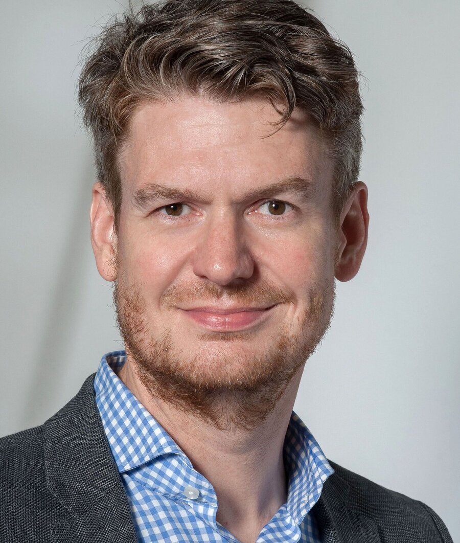 Portrait photo of a man with curly, dark blond hair and a beard, wearing a blue-and-white checked shirt and a grey blazer, smiling at the camera against a neutral light grey background.