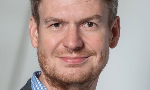 Porträtfoto eines Mannes mit lockigem, dunkelblondem Haar und Bart, der ein blau-weiß kariertes Hemd und ein graues Sakko trägt und lächelnd in die Kamera blickt, vor neutralem hellgrauem Hintergrund.