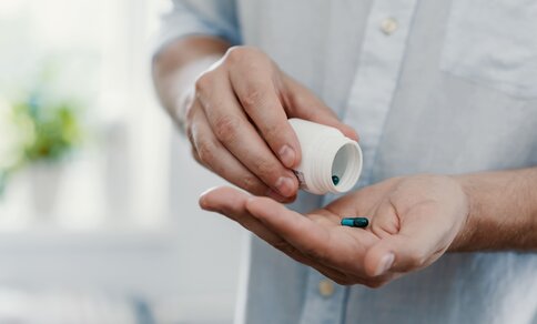 Nahaufnahme einer Person in hellgrauem Hemd, die eine kleine weiße Medikamentenflasche hält und Tabletten in die geöffnete Hand gibt.