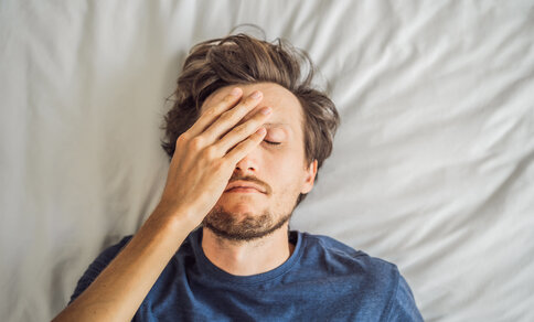 Ein Mann liegt mit geschlossenen Augen auf dem Rücken im Bett und legt sich erschöpft die Hand auf die Stirn. Er trägt ein blaues T-Shirt.